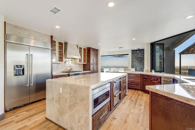 kitchen with stainless steel built in fridge, tasteful backsplash, a center island, wall chimney range hood, and light hardwood / wood-style floors