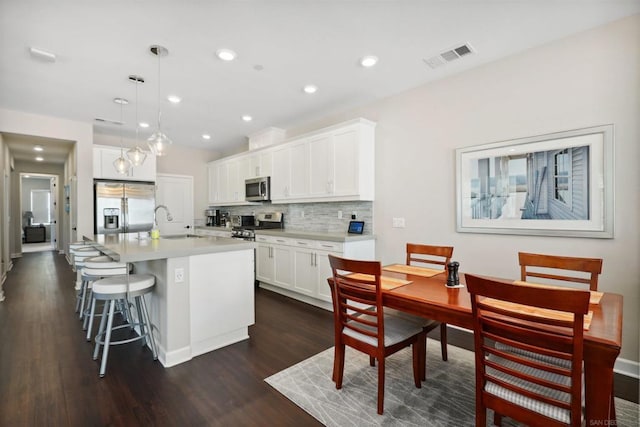 kitchen with dark wood-type flooring, hanging light fixtures, an island with sink, appliances with stainless steel finishes, and white cabinetry