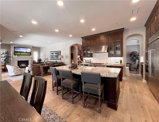 kitchen featuring a kitchen bar, light wood-type flooring, light stone counters, dark brown cabinets, and an island with sink