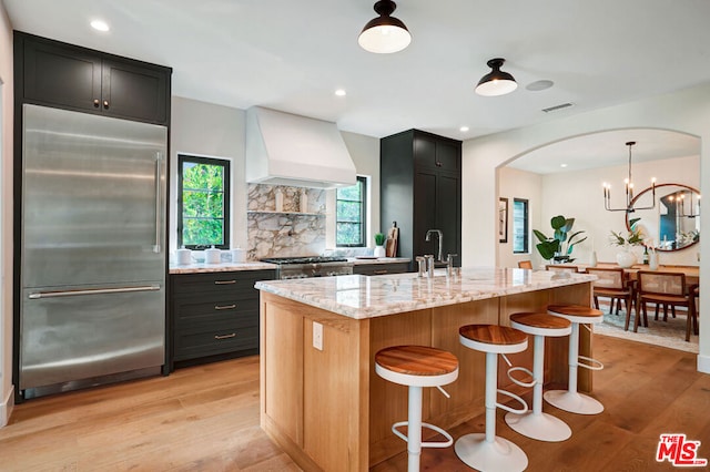 kitchen with light stone countertops, light hardwood / wood-style floors, a center island with sink, custom range hood, and appliances with stainless steel finishes