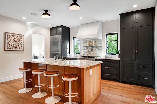 kitchen with light hardwood / wood-style floors, custom range hood, a kitchen island with sink, and appliances with stainless steel finishes