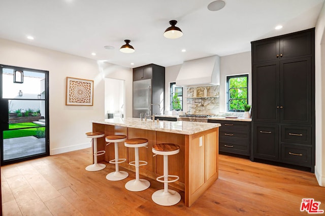 kitchen with custom exhaust hood, a kitchen island with sink, light stone counters, a kitchen bar, and stainless steel appliances