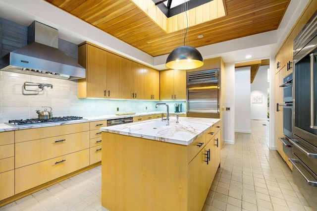 kitchen featuring wall chimney exhaust hood, sink, appliances with stainless steel finishes, pendant lighting, and a kitchen island with sink