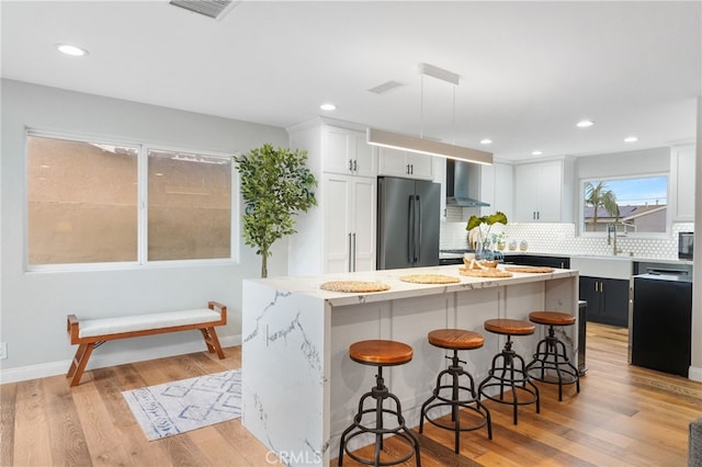 kitchen featuring wall chimney range hood, white cabinetry, a center island, light hardwood / wood-style floors, and stainless steel refrigerator