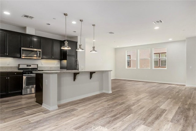 kitchen with light hardwood / wood-style floors, stainless steel appliances, hanging light fixtures, and an island with sink