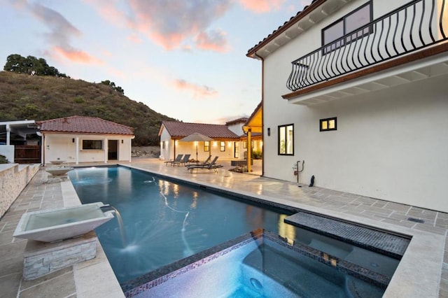 pool at dusk featuring a patio, an outdoor structure, and an in ground hot tub