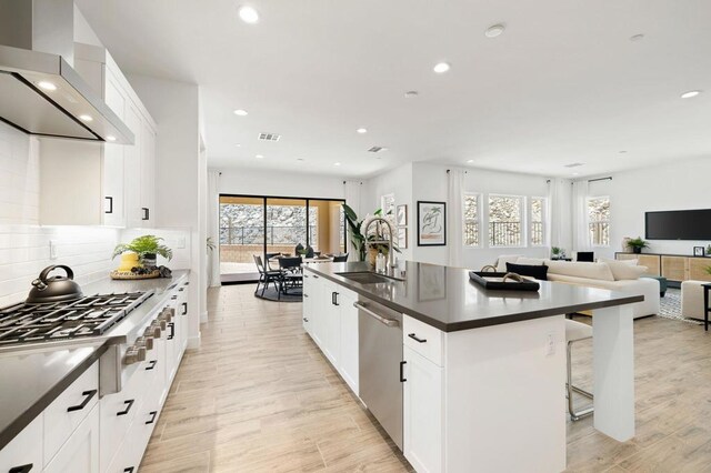 kitchen with appliances with stainless steel finishes, light wood-type flooring, wall chimney exhaust hood, a center island with sink, and white cabinetry