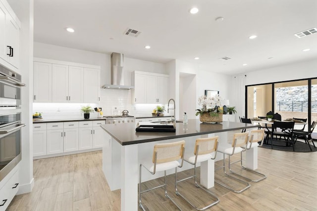 kitchen featuring a center island with sink, white cabinetry, a kitchen bar, and wall chimney range hood