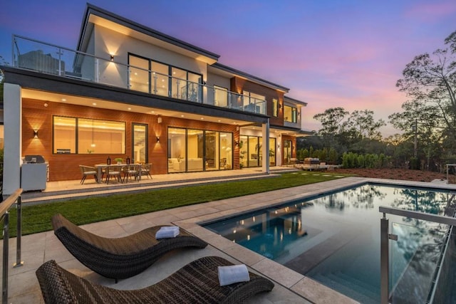 back house at dusk featuring a pool with hot tub, a balcony, and a patio