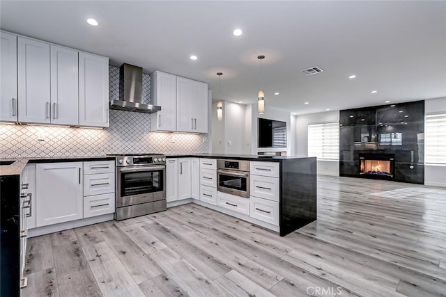 kitchen with white cabinets, wall chimney exhaust hood, light wood-type flooring, appliances with stainless steel finishes, and decorative light fixtures