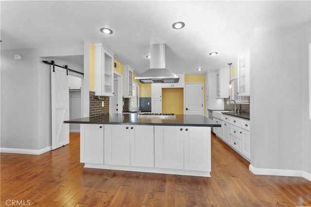 kitchen with a barn door, a peninsula, island exhaust hood, dark countertops, and white cabinets