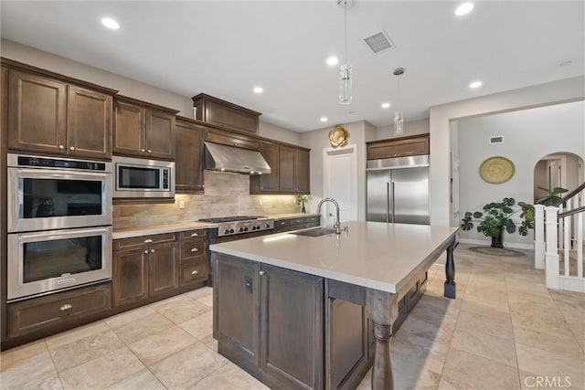 kitchen featuring tasteful backsplash, sink, wall chimney range hood, built in appliances, and decorative light fixtures