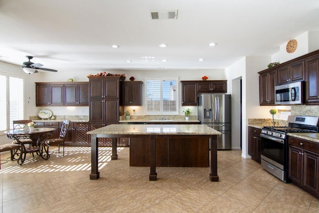 kitchen with a center island, ceiling fan, light stone countertops, appliances with stainless steel finishes, and dark brown cabinets