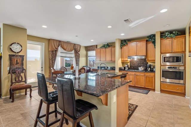 kitchen featuring stainless steel appliances, a kitchen island with sink, sink, dark stone countertops, and light tile patterned flooring