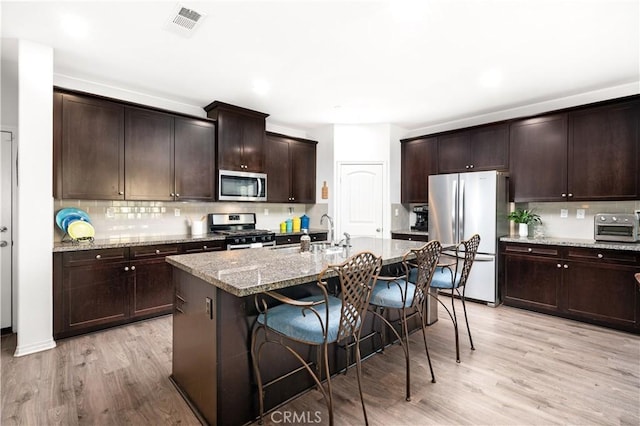 kitchen with sink, light stone counters, stainless steel appliances, dark brown cabinets, and a center island with sink