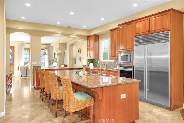 kitchen featuring a center island with sink, kitchen peninsula, a kitchen bar, built in appliances, and light stone counters