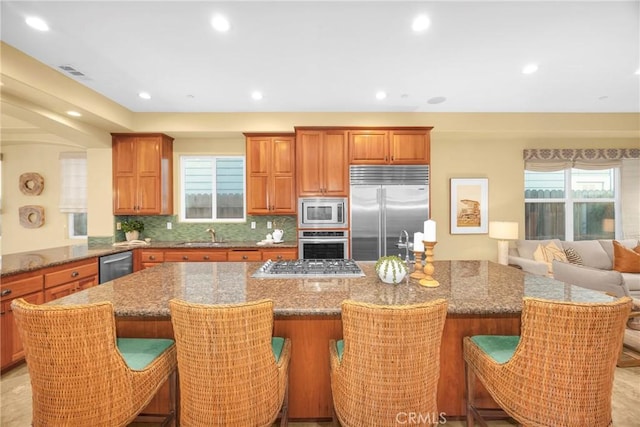 kitchen featuring a kitchen bar, backsplash, light stone countertops, built in appliances, and a center island