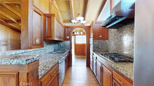 kitchen featuring brown cabinetry, arched walkways, a sink, appliances with stainless steel finishes, and wall chimney exhaust hood