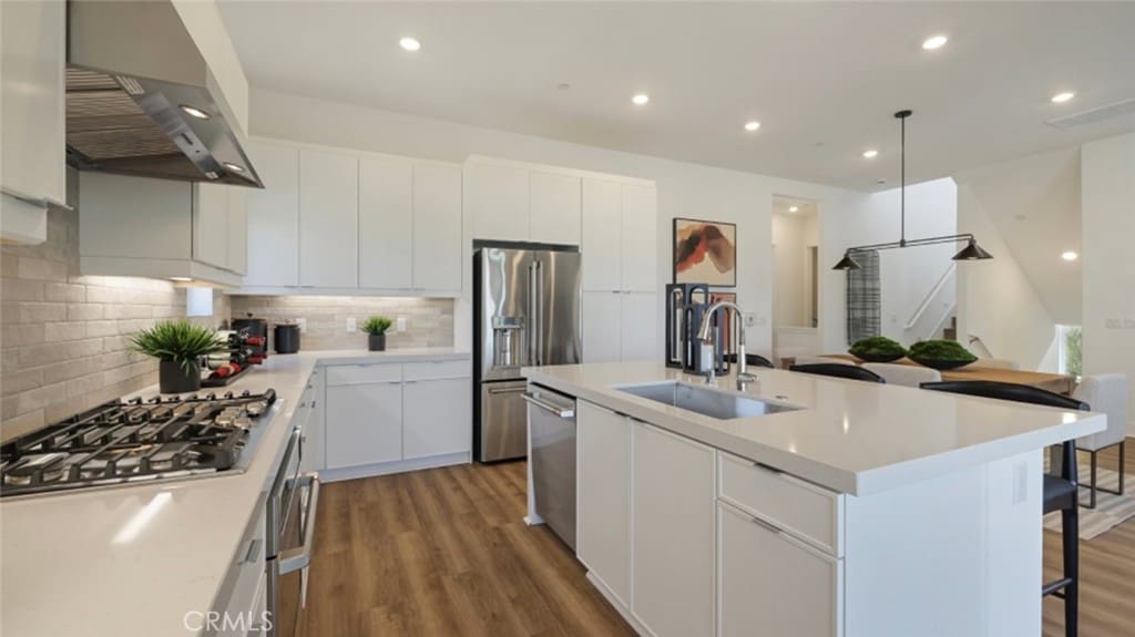 kitchen featuring appliances with stainless steel finishes, wall chimney exhaust hood, sink, a center island with sink, and white cabinetry