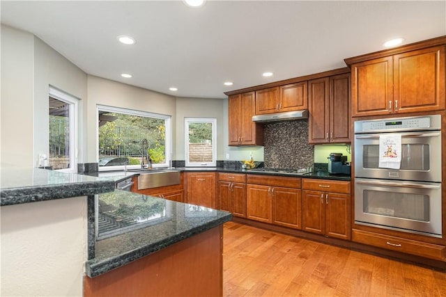 kitchen featuring decorative backsplash, stainless steel double oven, sink, light hardwood / wood-style flooring, and dark stone countertops