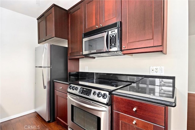 kitchen with reddish brown cabinets, dark wood-style flooring, dark countertops, appliances with stainless steel finishes, and baseboards