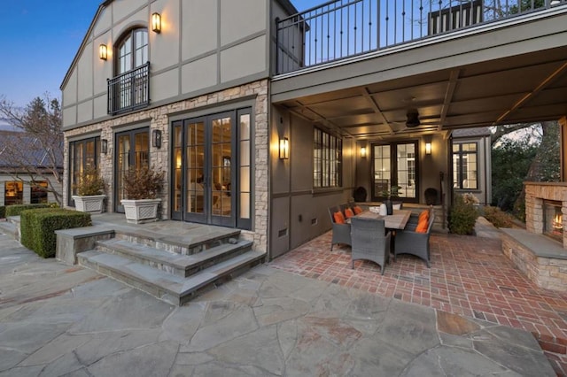 patio terrace at dusk featuring exterior fireplace, a balcony, french doors, and ceiling fan