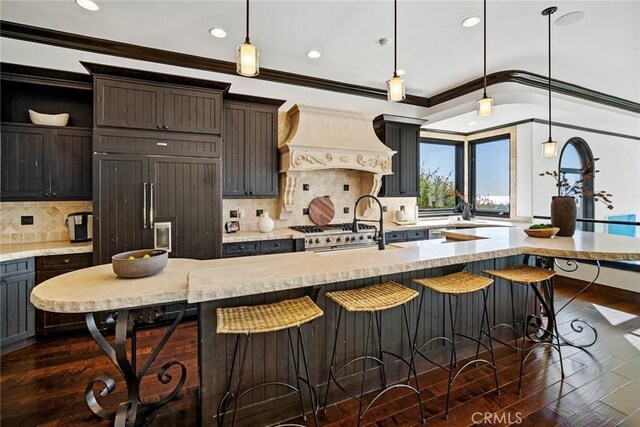 kitchen with decorative backsplash, custom range hood, a kitchen island with sink, dark hardwood / wood-style floors, and hanging light fixtures