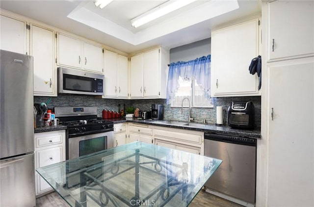 kitchen with decorative backsplash, appliances with stainless steel finishes, a raised ceiling, sink, and light hardwood / wood-style flooring