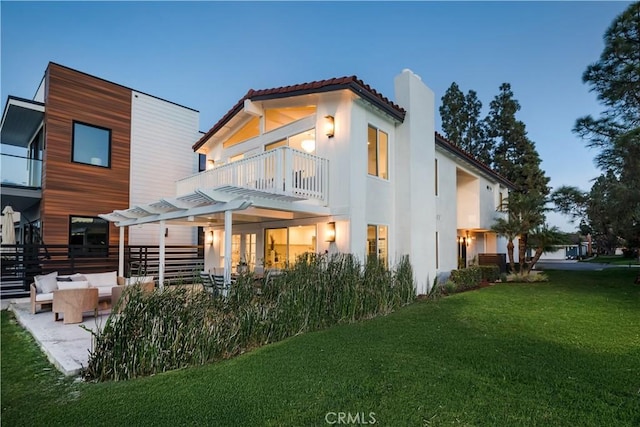 back house at dusk featuring a pergola, a balcony, outdoor lounge area, a patio area, and a lawn