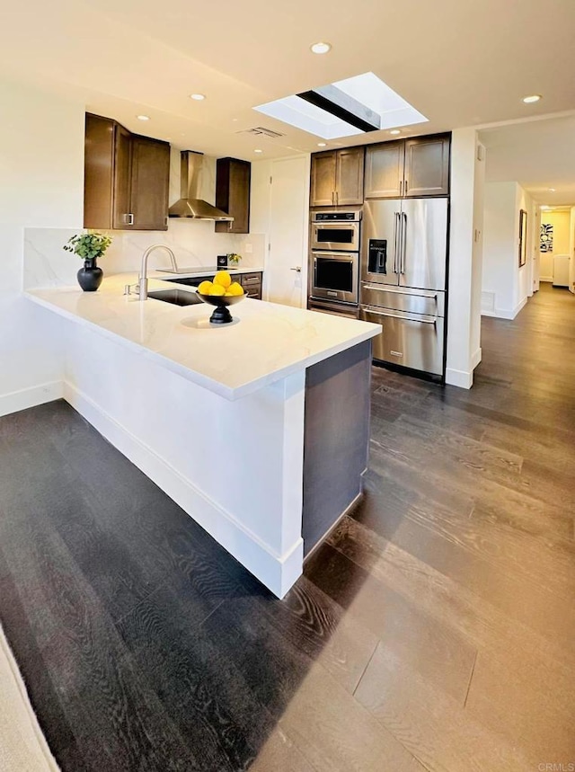 kitchen featuring a peninsula, a sink, light countertops, appliances with stainless steel finishes, and wall chimney exhaust hood