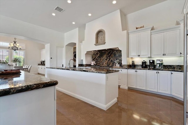 kitchen with white cabinets, an inviting chandelier, dark stone counters, and an island with sink