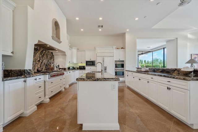 kitchen featuring white cabinetry, a center island with sink, built in appliances, and dark stone countertops