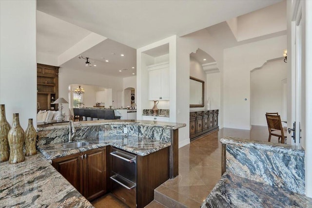 kitchen featuring kitchen peninsula, sink, dark brown cabinetry, light stone counters, and a chandelier