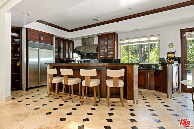 kitchen with a center island, wall chimney exhaust hood, built in fridge, a kitchen bar, and ornamental molding