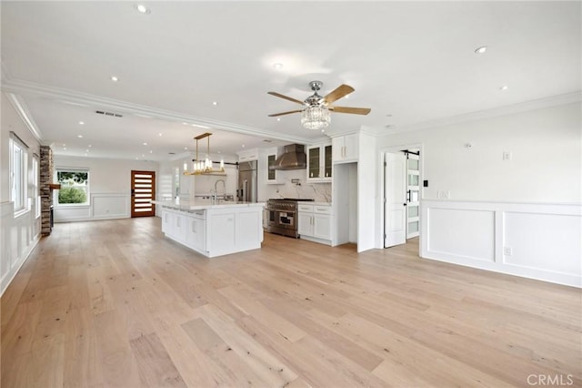 kitchen featuring high quality appliances, a center island with sink, white cabinets, wall chimney range hood, and light hardwood / wood-style flooring