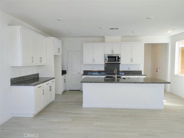 kitchen featuring an island with sink, white cabinets, dark stone counters, and light wood-type flooring