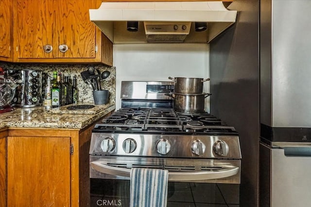 kitchen featuring stone counters, stainless steel gas range, range hood, tasteful backsplash, and brown cabinetry