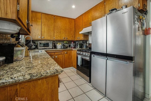 kitchen featuring light tile patterned floors, appliances with stainless steel finishes, brown cabinets, under cabinet range hood, and backsplash