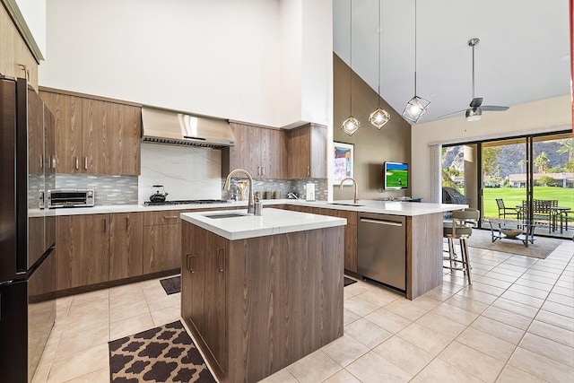 kitchen with ceiling fan, stainless steel appliances, wall chimney range hood, a towering ceiling, and an island with sink