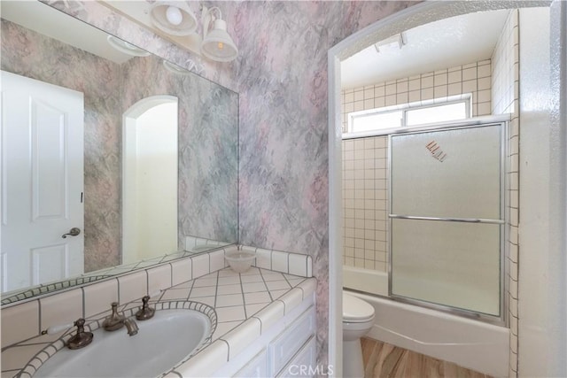 full bathroom featuring combined bath / shower with glass door, toilet, vanity, and wallpapered walls