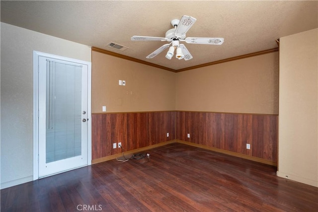 empty room featuring wooden walls, a ceiling fan, wood finished floors, visible vents, and wainscoting