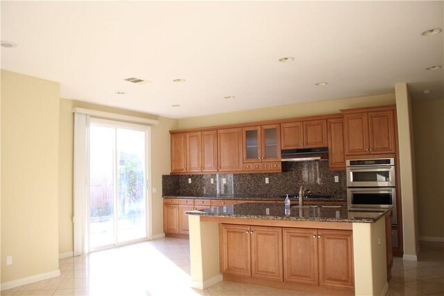 kitchen with light tile patterned floors, double oven, dark stone counters, and a center island with sink