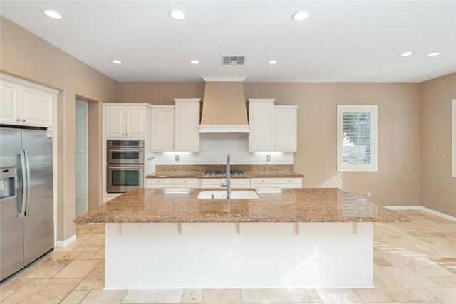 kitchen featuring light stone counters, custom range hood, stainless steel appliances, and an island with sink