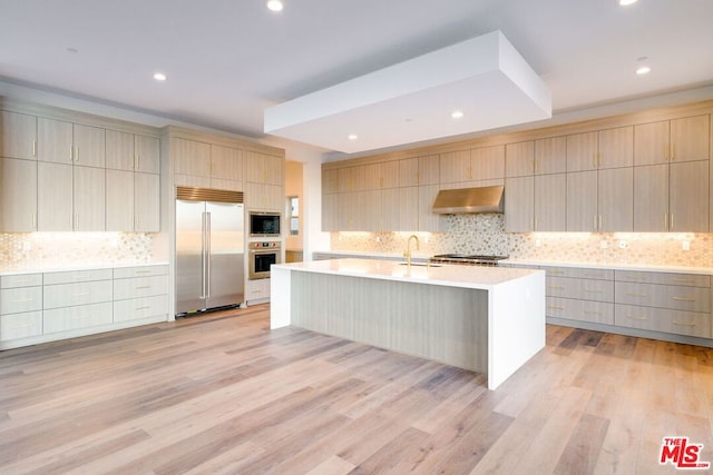 kitchen featuring light wood-type flooring, ventilation hood, a center island with sink, and built in appliances