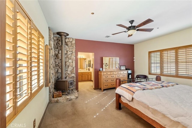 bedroom with a wood stove, ceiling fan, ensuite bath, and light colored carpet