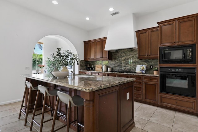 kitchen with premium range hood, dark stone counters, black appliances, a center island with sink, and sink