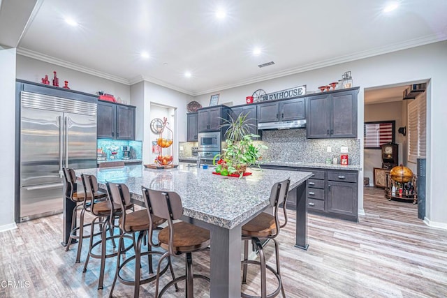 kitchen featuring a breakfast bar, a kitchen island with sink, built in appliances, light stone countertops, and light wood-type flooring