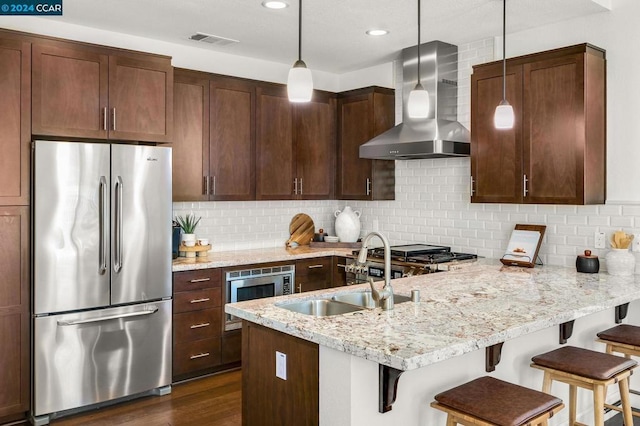 kitchen with stainless steel appliances, hanging light fixtures, light stone countertops, wall chimney exhaust hood, and sink