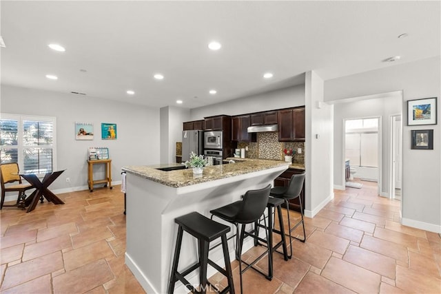 kitchen featuring appliances with stainless steel finishes, a breakfast bar, tasteful backsplash, light stone counters, and a center island with sink