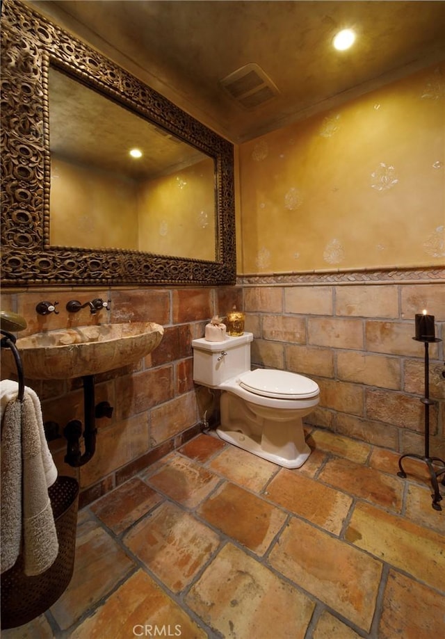half bathroom featuring stone tile floors, visible vents, toilet, tile walls, and wainscoting